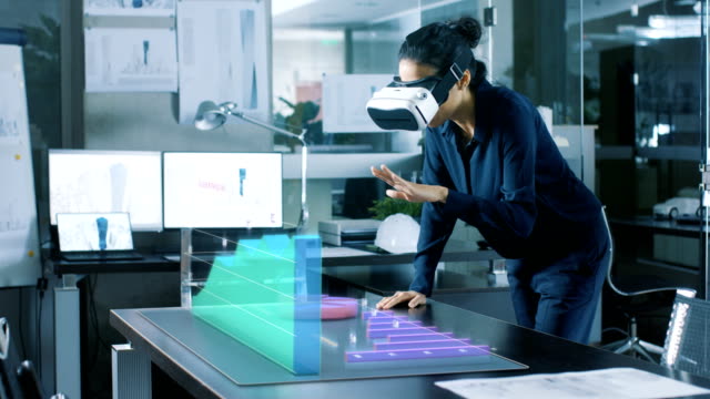 Die-Office-berufstätige-Frau-interagiert-tragen-Augmented-Reality-Kopfhörer-mit-Infografik-zeigt-Statistiken.--Sie-lehnt-sich-auf-dem-Tisch-mit-animierten-3D-Modelle-zeigen-Wachstum-des-Unternehmens.