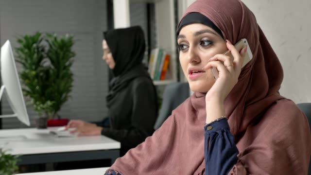 Joven-hermosa-chica-en-hijab-rosa-se-sienta-en-la-oficina-y-habla-en-el-teléfono-inteligente,-riendo.-60-fps