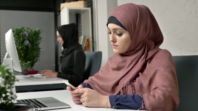 Young-beautiful-girl-in-pink-hijab-sits-in-office-and-uses-smartphone.-Girl-in-black-hijab-in-the-background.-Arab-women-in-the-office.-60-fps