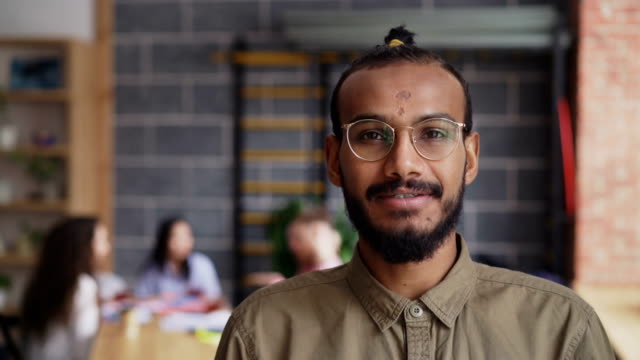 Young-african-american-male-entrepreneur-in-shirt-and-glasses-smiling-and-looking-at-camera-while-his-colleagues-working-in-modern-office-indoors