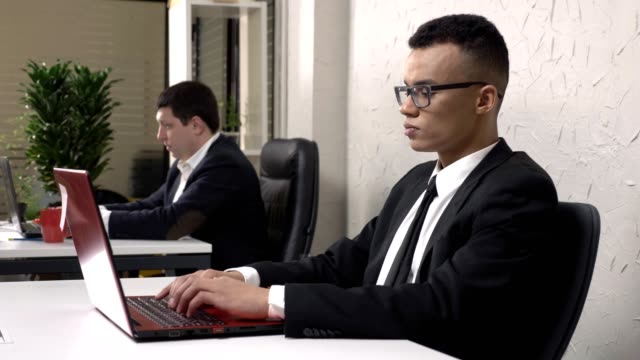 Young-successful-African-businessman-wearing-glasses-is-sitting-in-the-office-and-working-on-a-laptop,-takes-off-his-glasses,-looks-into-the-camera,-smiles-and-shakes-his-head,-Caucasian-man-in-a-suit-in-the-background.-60-fps