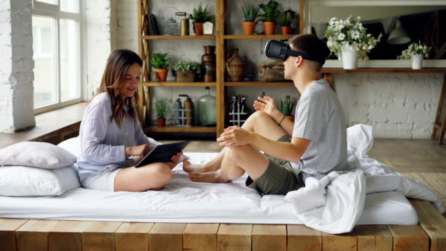 Cute-couple-husband-and-wife-are-having-fun-with-augmented-reality-glasses,-man-is-wearing-glasses-and-moving-hands,-woman-is-using-tablet-and-laughing.
