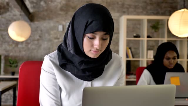 Retratos-de-mujeres-jóvenes-musulmanas-en-hijab-trabajando-y-mirando-en-la-cámara,-sonriendo,-dos-mujeres-sentadas-en-la-oficina-moderna
