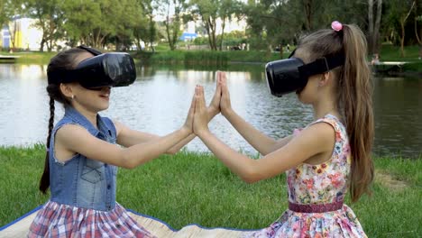 Zwei-kleine-Mädchen-in-virtual-Reality-Brille-sitzen-und-spielen-im-Park-auf-dem-Rasen-bei-Sonnenuntergang-gegen-den-Fluss.-Sie-halten-einander-die-Hände-und-lachen.