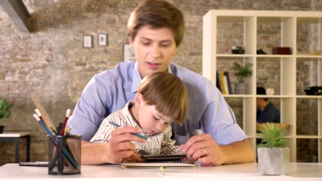Joven-padre-sosteniendo-a-su-pequeño-hijo-y-de-la-escritura-sobre-papel,-chico-con-tableta,-sentado-en-la-oficina-moderna