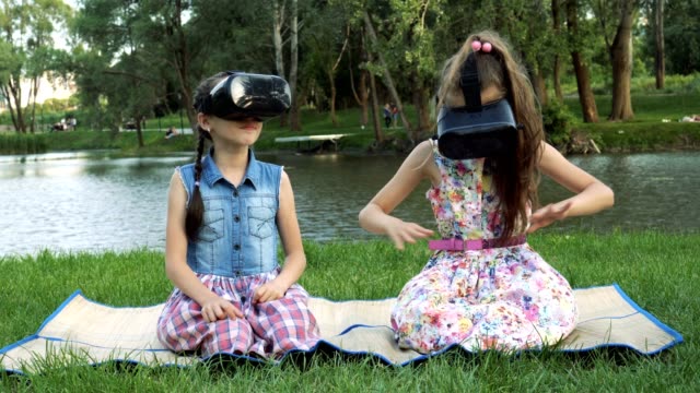 Two-little-girls-play-virtual-games-in-the-Park-on-the-river-Bank.-They-sit-in-virtual-reality-glasses,-look-around-and-wave-their-hands.