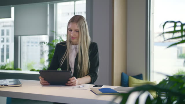 Stylish-business-woman-with-tablet-in-office.-Elegant-woman-in-suit-sitting-at-table-with-laptop-and-surfing-tablet-in-modern-light-office-with-big-windows