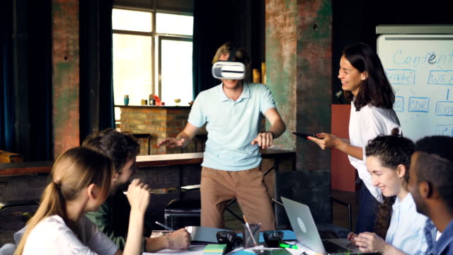 Junger-Mann-mit-virtual-Reality-Brille-ist-Spaß-Spiel-spielen,-während-seine-Kollegen-ihn-ansah,-lachen-und-Verwendung-von-Notebook-und-Tablet-in-Office.