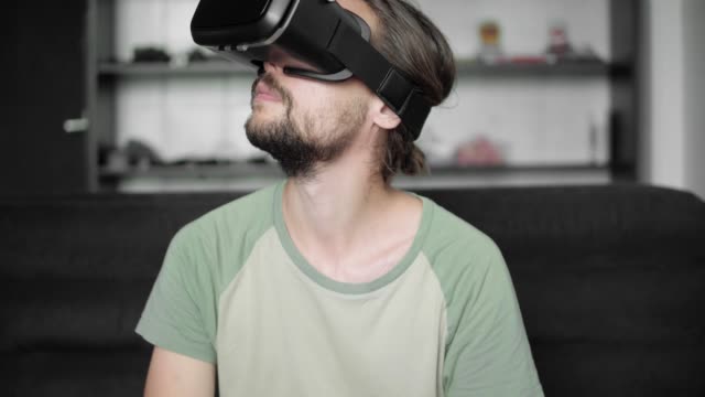 Young-bearded-hipster-man-using-his-VR-headset-display-with-headphones-for-virtual-reality-game-or-watching-the-360-video-while-sitting-on-sofa-at-home-in-the-living-room.-VR-Technology.