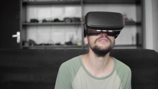 Joven-barbudo-a-hombre-hipster-usando-su-pantalla-del-auricular-VR-para-juego-de-realidad-virtual-o-ver-el-video-360-mientras-está-sentado-en-el-sofá-en-casa-en-la-sala-de-estar.-Tecnología-VR.