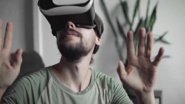 Young-bearded-hipster-man-using-his-VR-headset-display-for-virtual-reality-game-or-watching-the-360-video-and-trying-to-touch-to-something-he-see-and-catch-while-sitting-on-sofa.-VR-Technology.