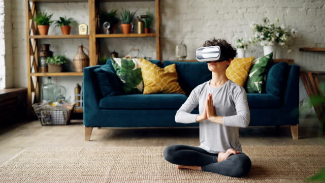 Slim-young-lady-in-artificial-reality-glasses-is-meditating-at-home-in-lotus-pose-with-hands-in-namaste-enjoying-simulation-and-relaxing.-Modern-technology-and-experience-concept.