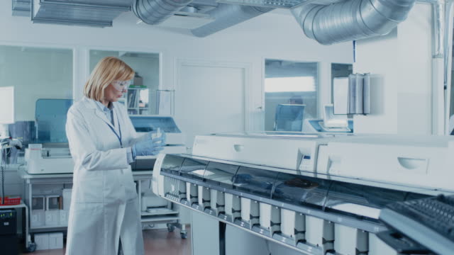 En-laboratorio-científico-femenino-toma-un-tubo-de-ensayo-con-muestras-de-sangre-saliendo-del-equipo-médico-y-analiza-los-datos-con-ordenador.-Equipo-de-investigadores-trabaja-en-laboratorio-farmacéutico.