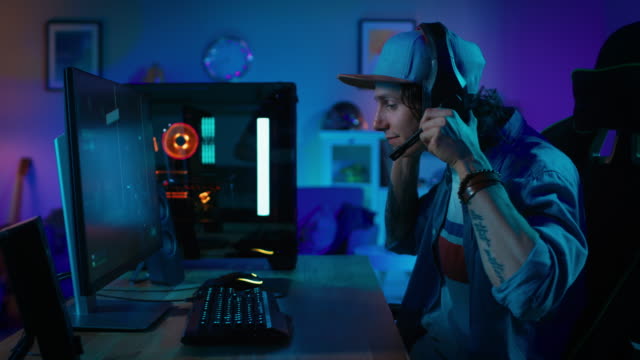 Professional-Gamer-Puts-His-Headset-with-a-Microphone-On-and-Starts-Playing-First-Person-Shooter-Online-Video-Game-on-His-Powerful-Personal-Computer-with-Colorful-Neon-Led-Lights.-Evening.