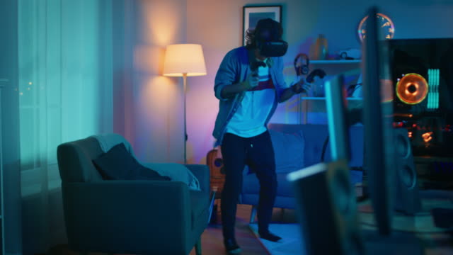 Young-Man-is-Energetically-Playing-a-Virtual-Reality-Video-Game-While-Wearing-a-Headset.-He's-is-Standing-in-a-Cozy-Room-with-Warm-Neon-Lights.-He's-Swinging-Hands,-Shooting-and-Dodging-Obstacles.