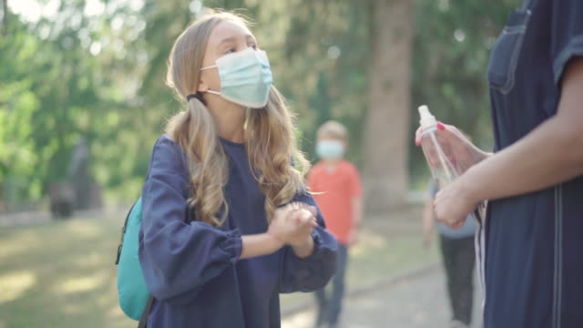 Unrecognizable-woman-spraying-sanitizer-on-hands-of-schoolgirl-at-schoolyard.-Cute-girl-in-face-mask-disinfecting-palms-and-running-away.-Education-during-Covid-19-pandemic.