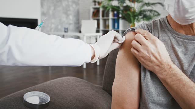 Ärztin-in-Einweghandschuhen-injiziert-Impfstoff-in-die-Hand-von-Senior-Männlich-in-Schutzmaske,-die-auf-der-Couch-zu-Hause-sitzen