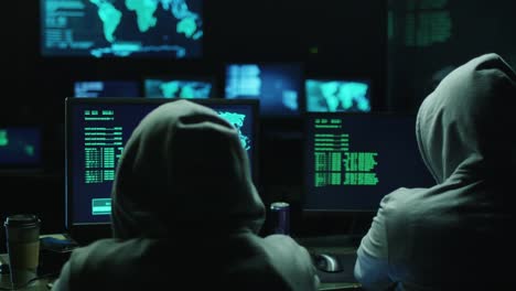 Dos-los-hakers-en-carcasas-trabajo-en-las-computadoras-con-mapas-y-datos-en-pantallas-de-oficina-en-una-sala-oscura.