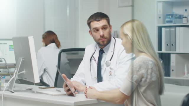 In-Doctor's-Office-Professional-Doctor-Consults-Beautiful-Young-Woman-with-a-Help-of-a-Tablet.-They-Look-and-the-Screen-and-Smile.