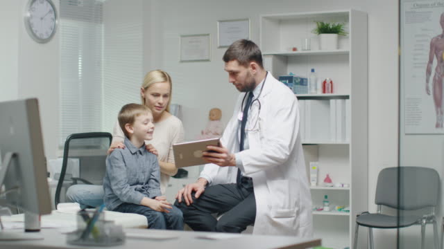 Male-Doctor-Consults-Young-Boy-and-His-Mother-with-a-Help-of-a-Tablet.-They-all-Smile.