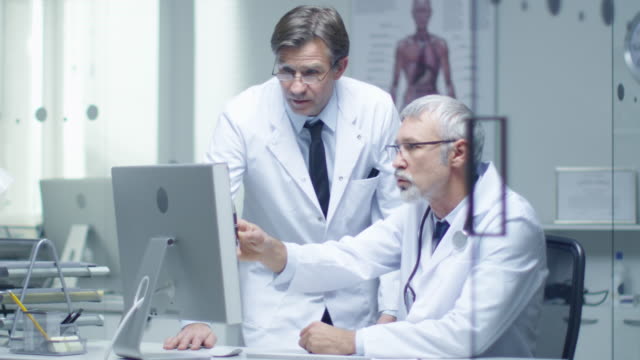 Senior-Doctor-and-His-Assistant-Discuss-Patient's-Log-on-Personal-Computer.-In-Slow-Motion.
