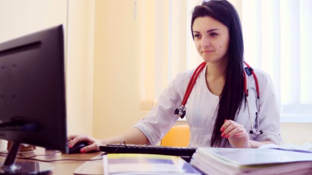 Young-female-doctor-with-stethoscope-sitting-at-the-table-near-the-window-and-typing