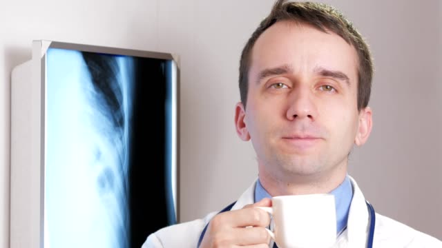 A-young-doctor-looking-at-the-camera-is-drinking-a-hot-drink-from-a-white-mug.-On-the-neck-hangs-a-stethoscope.-Close-up