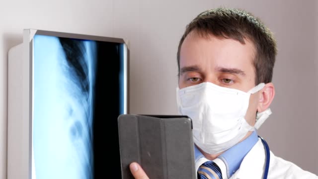 A-young-doctor-in-a-face-mask-uses-a-tablet-computer-in-the-clinic.-A-man-records-the-results-of-a-patient's-x-ray