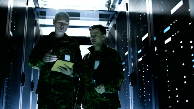 Two-Soldiers-Walking-in-Data-Center-Corridor.-One-Holds-Tablet-Computer,-They-Have-Discussion.-Rows-of-Working-Data-Servers-by-their-Sides.