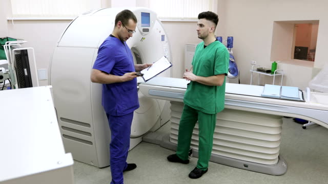 Zwei-Ärzte-sind-in-einem-Raum-mit-einem-CT-Scanner.-Der-junge-Spezialist-berät-über-Mobiltelefon.
