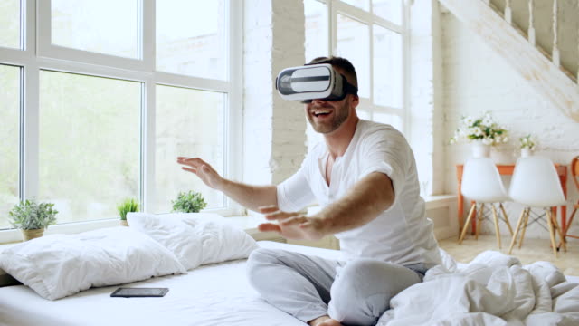 Young-cheerful-man-wearing-virtual-reality-headset-having-360-VR-video-experience-while-sitting-in-bed-at-home