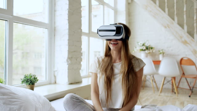 Mujer-alegre-joven-con-casco-de-realidad-virtual-ver-360-VR-video-película-sentado-en-la-cama-en-casa