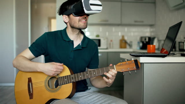 Feliz-joven-sentado-en-la-cocina-aprendiendo-a-tocar-la-guitarra-usando-VR-360-auriculares-y-se-le-siente-guitarrista-en-concierto-en-casa