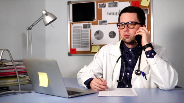 Männlichen-Arzt-in-der-Klinik-am-Schreibtisch-sitzen-und-reden-von-Smartphone.