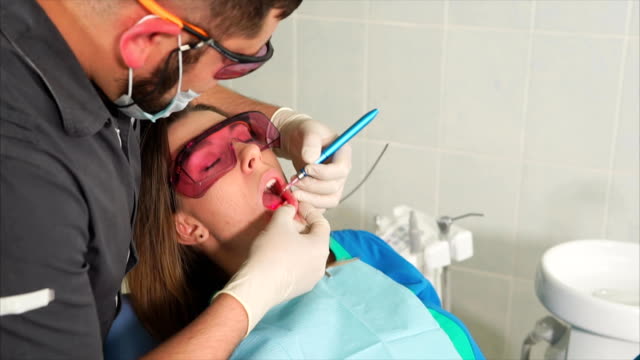 Doctor-orthodontist-with-glasses-whitens-the-teeth-of-a-young-woman-with-a-laser