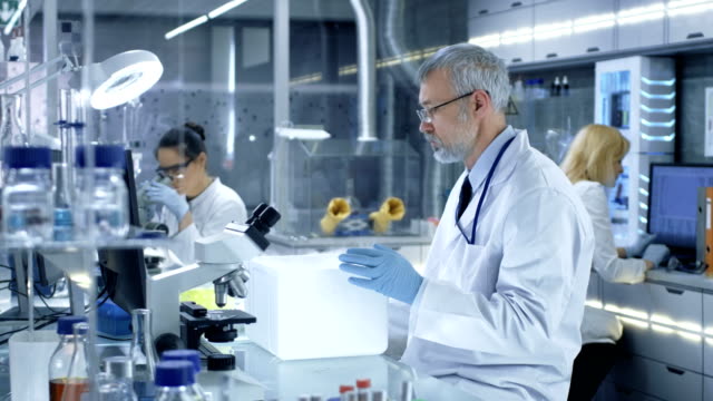Senior-Medical-Research-Scientist-Opens-Refrigerator-Box-Takes-Out-Petri-Dish-with-Samples-and-Examines-it.-He-Works-in-a-Busy-Modern-Laboratory-Center.