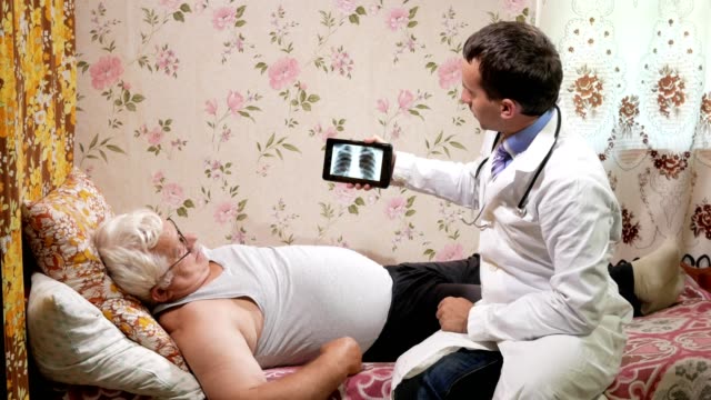 Un-médico-hombre-visita-a-un-paciente-en-casa.-Muestra-los-resultados-de-la-radiografía-en-un-equipo-tablet-PC.-El-hombre-está-acostado-en-el-sofá