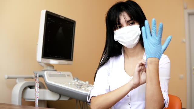Atractiva-doctora-sonriente-en-guantes-médicos-de-vestirse-de-máscara-quirúrgica-sentado-en-la-mesa-de-su-oficina.-Rodada-en-4k