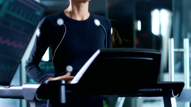 Schöne-Frau-Athlet-mit-Elektroden-angeschlossen-an-ihren-Körper-läuft-auf-einem-Laufband-in-einem-Sport-Wissenschaft-Labor.-Im-Hintergrund-Hightech-Labor-mit-Monitore-mit-EKG-Messungen.