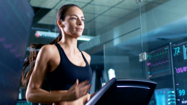 In-wissenschaftlichen-Sport-Labor,-die-schöne-Sportlerin-auf-einem-Laufband-mit-Elektroden-an-ihrem-Körper-geht,-anzeigen-Monitore-EKG-Daten-auf-dem-Display.-Slow-Motion.-Niedrigen-Winkel.