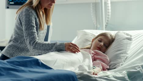 Cute-Little-Girl-Sleeps-on-a-Bed-in-the-Children's-Hospital,-Caring-Mother-Covers-Her-with-a-Blanket-and-Caresses-Her-Forehead.-Modern-Pediatric-Ward.