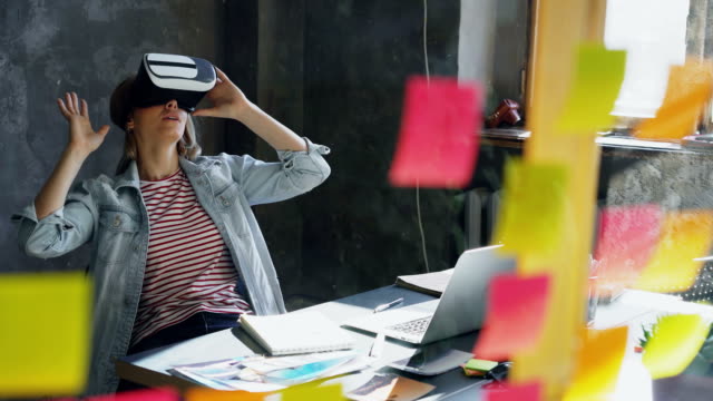 Attraktive-junge-Frau-sitzt-am-Tisch-und-virtual-Reality-Brille.-Sie-bewegt-sich-Hände-und-und-mit-VR-Erlebnis-in-modernen-Lof-Büro