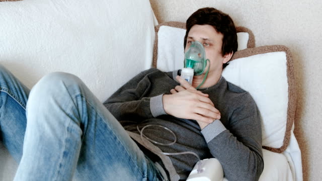Verwenden-Sie-Vernebler-und-Inhalator-für-die-Behandlung.-Junger-Mann-Einatmen-durch-Inhalator-Maske-auf-der-Couch-liegen.-Vorderansicht