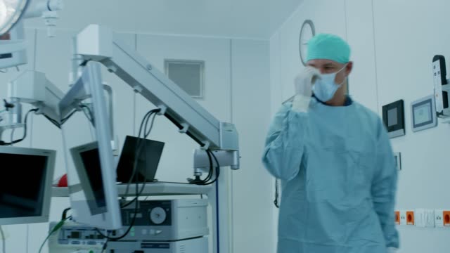 Senior-Surgeon-Enters-Operating-Room-Where-His-Assistants-and-Patient-Wait,-He-Puts-on-a-Mask-and-Starts-Surgery.-Real-Modern-Hospital-with-Authentic-Equipment.