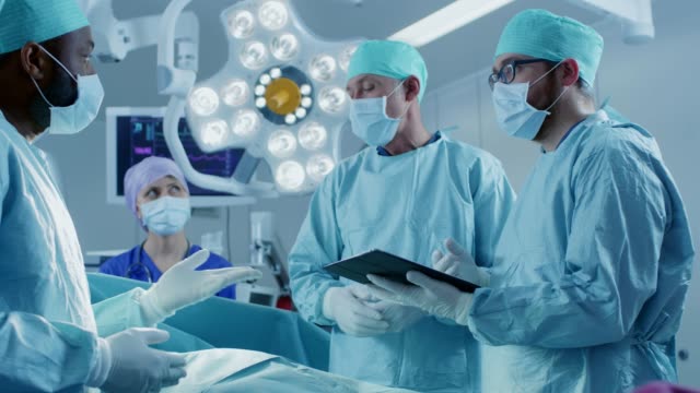 Professionelle-Chirurgen-und-Assistenten-sprechen-und-digitale-Tablettcomputer-verwenden,-während-der-Operation.-Sie-arbeiten-im-modernen-Krankenhaus-OP-Saal.