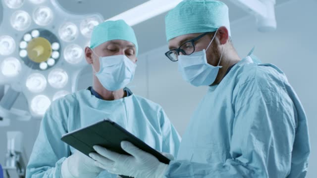 Dos-profesionales-cirujanos-uso-Digital-Tablet-PC-mientras-permanente-en-la-sala-de-operaciones-moderna-del-Hospital.
