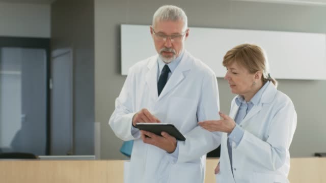 In-the-Hospital-Doctors-Walking-through-Lobby,-have-Discussion-while-Using-Tablet-Computer.-In-the-Background-Patients-and-Medical-Personnel.-New-Modern-Fully-Functional-Medical-Facility.