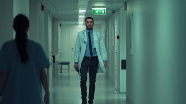 Determined-Handsome-Doctor-Wearing-White-Coat-with-Stethoscope-Walks-Through-Hospital-Hallway-in-Slow-motion.-Modern-Bright-Clinic-with-Professional-Staff.