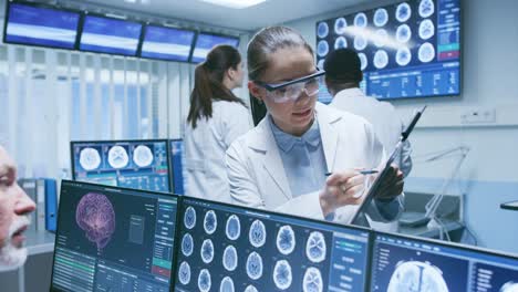 Senior-Medical-Scientist-Consults-Female-Apprentice-with-Clipboard,-He-Works-with-CT-Brain-Scan-Images-on-a-Personal-Computer-in-Laboratory.-Neurologists-in-Research-Center-Work-on-Brain-Tumor-Cure.