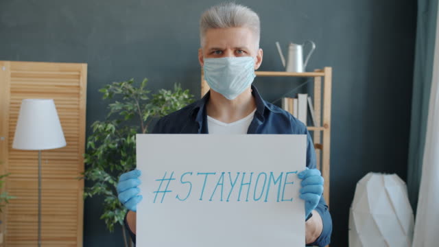 Slow-motion-portrait-of-adult-man-wearing-mask-and-gloves-holding-stayhome-poster-at-home